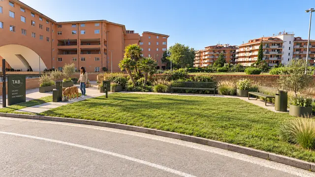 Il giardino per i cani fuori da TAB: il prato con le panchine, una ragazza col suo cane alla ciotola dell'acqua e dietro i palazzi di mattoni arancioni dell'ex Saint Gobain.