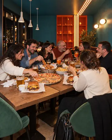 Sette amici a tavola la sera, con la pizza, un cestello di pollo fritto e due smash burger sullo stesso tavolo.